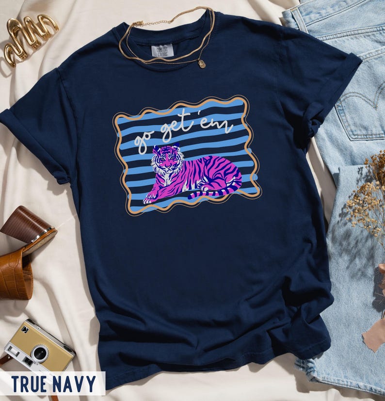 Pu&ograve; includere: T-shirt blu navy con grafica di tigre rosa e viola con la scritta "go get 'em" in una cornice blu e oro. La maglietta &egrave; in stile casual.