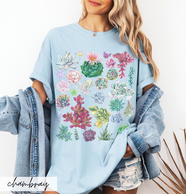 Puede incluir: Una camiseta azul claro con un estampado de suculentas coloridas. La camiseta presenta una variedad de suculentas en diferentes tonos de verde, rosa y rojo.