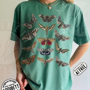 May include: A light green short-sleeve t-shirt featuring a graphic print of twelve various moth species in shades of brown, gray, and red. The shirt has a crew neckline and a relaxed fit. A gold-toned chain necklace is worn around the neck.