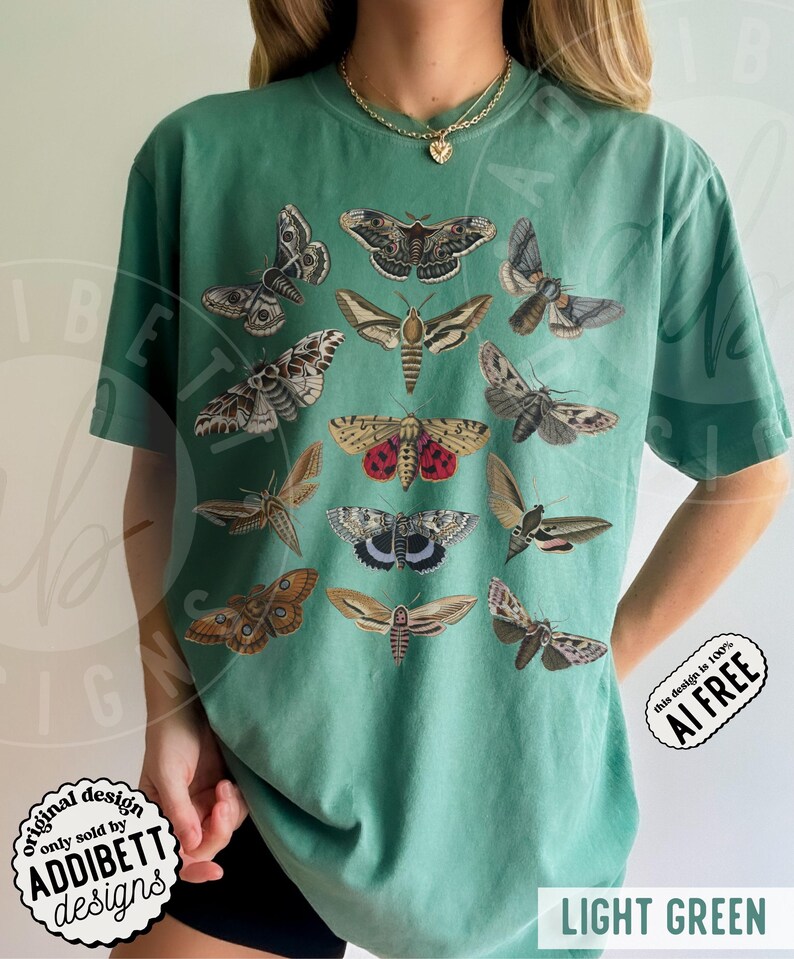 May include: A light green short-sleeve t-shirt featuring a graphic print of twelve various moth species in shades of brown, gray, and red. The shirt has a crew neckline and a relaxed fit. A gold-toned chain necklace is worn around the neck.