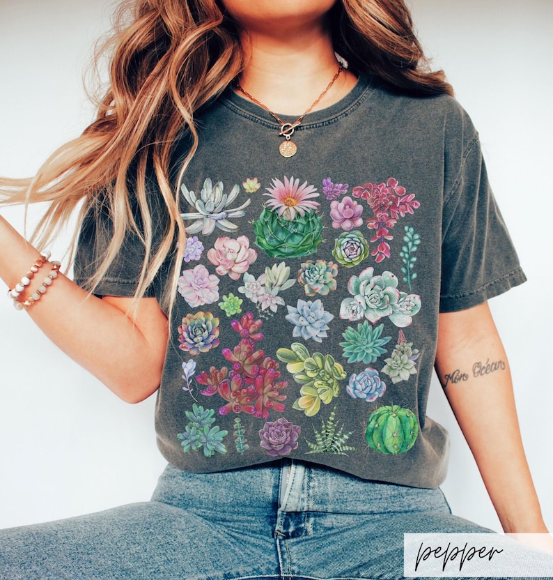 Puede incluir: Una camiseta gris oscuro con un estampado de suculentas coloridas. El estampado presenta una variedad de suculentas en tonos de verde, rosa, morado y rojo.