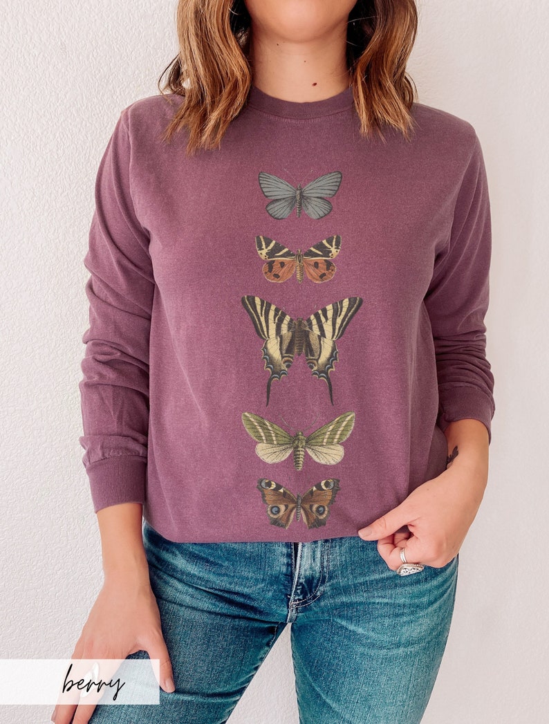 May include: A long-sleeved, purple sweatshirt with a vertical row of five different butterfly illustrations printed on the front. The butterflies are in various colors and patterns, including blue, brown, yellow, and black.
