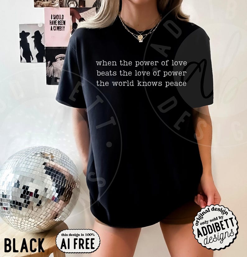 May include: A black short-sleeved t-shirt featuring white text that reads, when the power of love beats the love of power the world knows peace. The shirt has a crew neckline and a relaxed fit, showcasing a simple design with a clear, legible message.