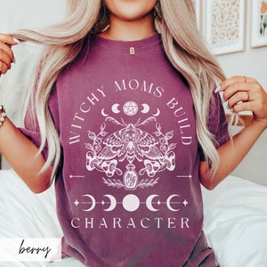 May include: A purple t-shirt with the text "Witchy Moms Build Character" in white. The design features a moth, a potion bottle, and a crescent moon with a pentagram.