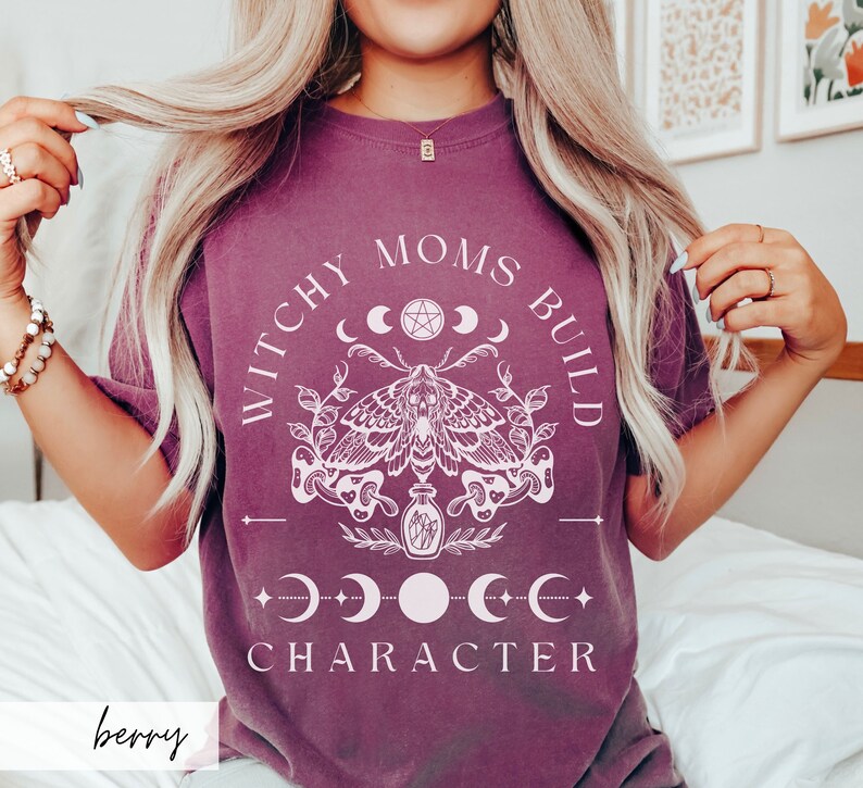 May include: A purple t-shirt with the text "Witchy Moms Build Character" in white. The design features a moth, a potion bottle, and a crescent moon with a pentagram.