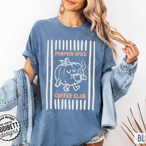 May include: A blue jean coloured t-shirt featuring a white graphic of a pumpkin character drinking from a mug. The text PUMPKIN SPICE COFFEE CLUB is printed in orange above and below the pumpkin. The design is framed by vertical white stripes.