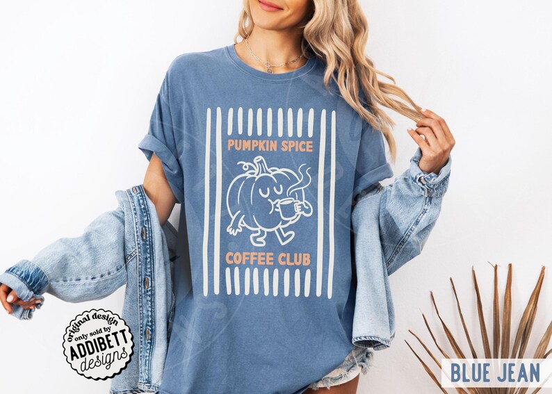 May include: A blue jean coloured t-shirt featuring a white graphic of a pumpkin character drinking from a mug. The text PUMPKIN SPICE COFFEE CLUB is printed in orange above and below the pumpkin. The design is framed by vertical white stripes.