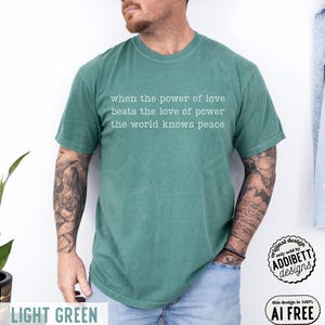 May include: A short-sleeved, light green t-shirt featuring white text printed on the chest that reads: when the power of love beats the love of power the world knows peace. The shirt has a crew neckline and a relaxed fit, shown in a solid color.