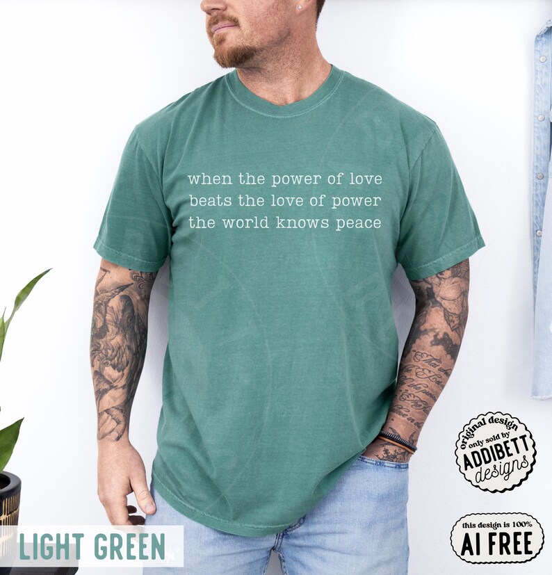 May include: A short-sleeved, light green t-shirt featuring white text printed on the chest that reads: when the power of love beats the love of power the world knows peace. The shirt has a crew neckline and a relaxed fit, shown in a solid color.