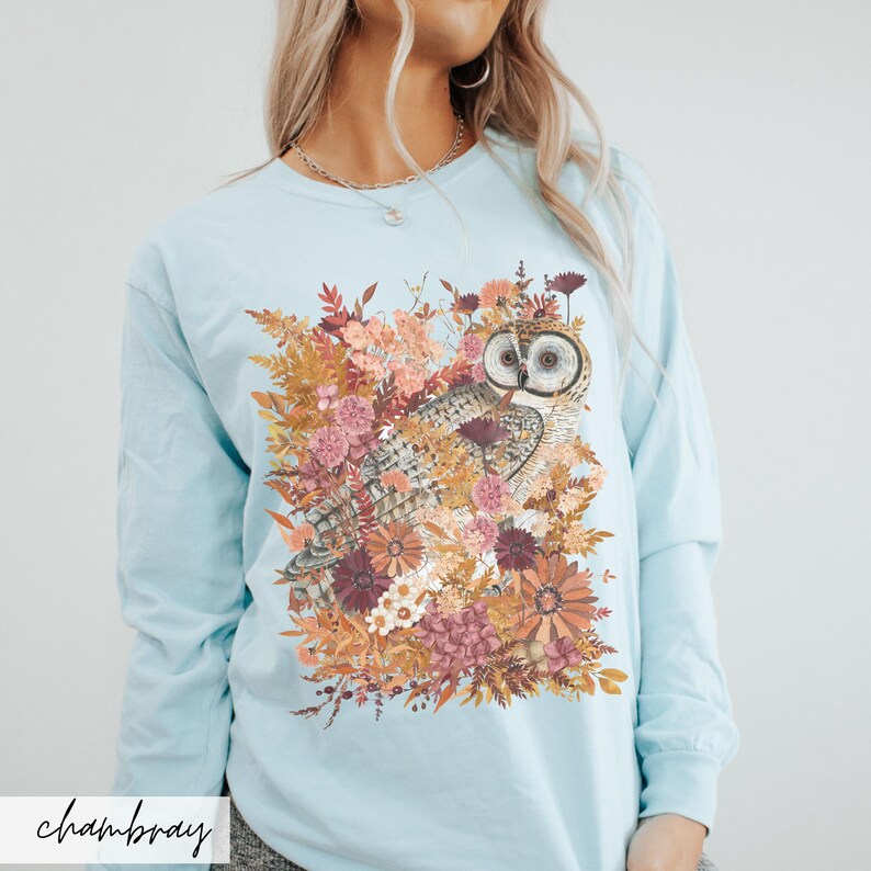 Pode incluir: Camiseta de manga comprida azul claro com uma estampa gr&aacute;fica de uma coruja cercada por flores e folhas de outono.