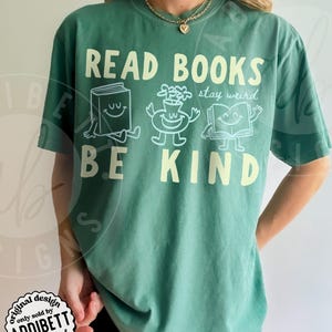 May include: A light green short-sleeve t-shirt featuring the text READ BOOKS BE KIND in large cream letters. Between the text are three white line-art illustrations of a smiling book, a smiling potted plant, and a smiling open book with the text stay weird.
