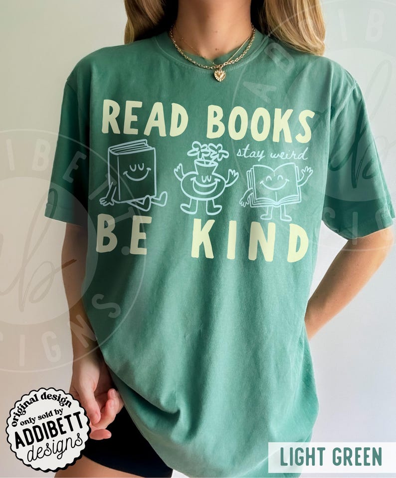 May include: A light green short-sleeve t-shirt featuring the text READ BOOKS BE KIND in large cream letters. Between the text are three white line-art illustrations of a smiling book, a smiling potted plant, and a smiling open book with the text stay weird.