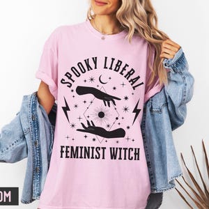 Puede incluir: Camiseta rosa claro con el texto negro "SPOOKY LIBERAL FEMINIST WITCH". El dise&ntilde;o incluye manos, estrellas, una luna creciente, rayos y un sol. La camiseta se lleva con una chaqueta vaquera.
