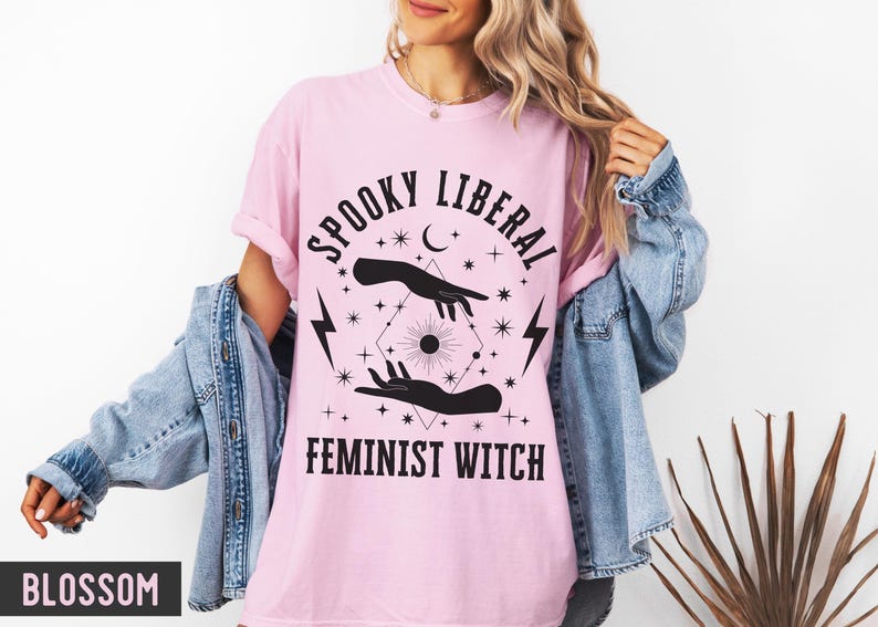 Puede incluir: Camiseta rosa claro con el texto negro "SPOOKY LIBERAL FEMINIST WITCH". El dise&ntilde;o incluye manos, estrellas, una luna creciente, rayos y un sol. La camiseta se lleva con una chaqueta vaquera.