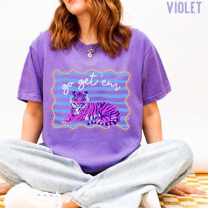 Pu&ograve; includere: T-shirt viola con un'illustrazione di tigre a strisce rosa e nere. La maglietta ha la scritta "go get 'em" in una cornice ondulata blu e oro. La maglietta &egrave; di colore viola unito.