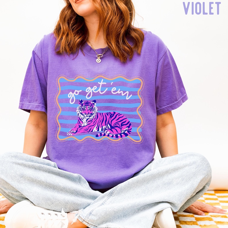 Pu&ograve; includere: T-shirt viola con un'illustrazione di tigre a strisce rosa e nere. La maglietta ha la scritta "go get 'em" in una cornice ondulata blu e oro. La maglietta &egrave; di colore viola unito.