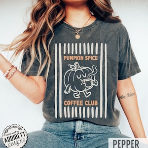 May include: A charcoal grey short-sleeve t-shirt featuring a graphic of a cartoon pumpkin holding a coffee mug. The text reads PUMPKIN SPICE COFFEE CLUB, framed by vertical white stripes. The design is printed in white and light orange ink.