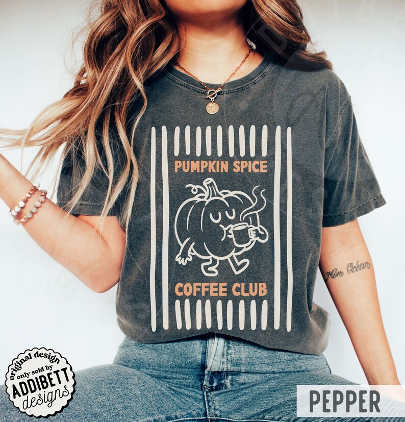 May include: A charcoal grey short-sleeve t-shirt featuring a graphic of a cartoon pumpkin holding a coffee mug. The text reads PUMPKIN SPICE COFFEE CLUB, framed by vertical white stripes. The design is printed in white and light orange ink.