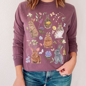 May include: A purple long-sleeved sweatshirt with a floral print featuring multiple bunnies in various poses. The bunnies are brown, white, and gray with pink noses and whiskers. The flowers are in shades of pink, purple, blue, and yellow.