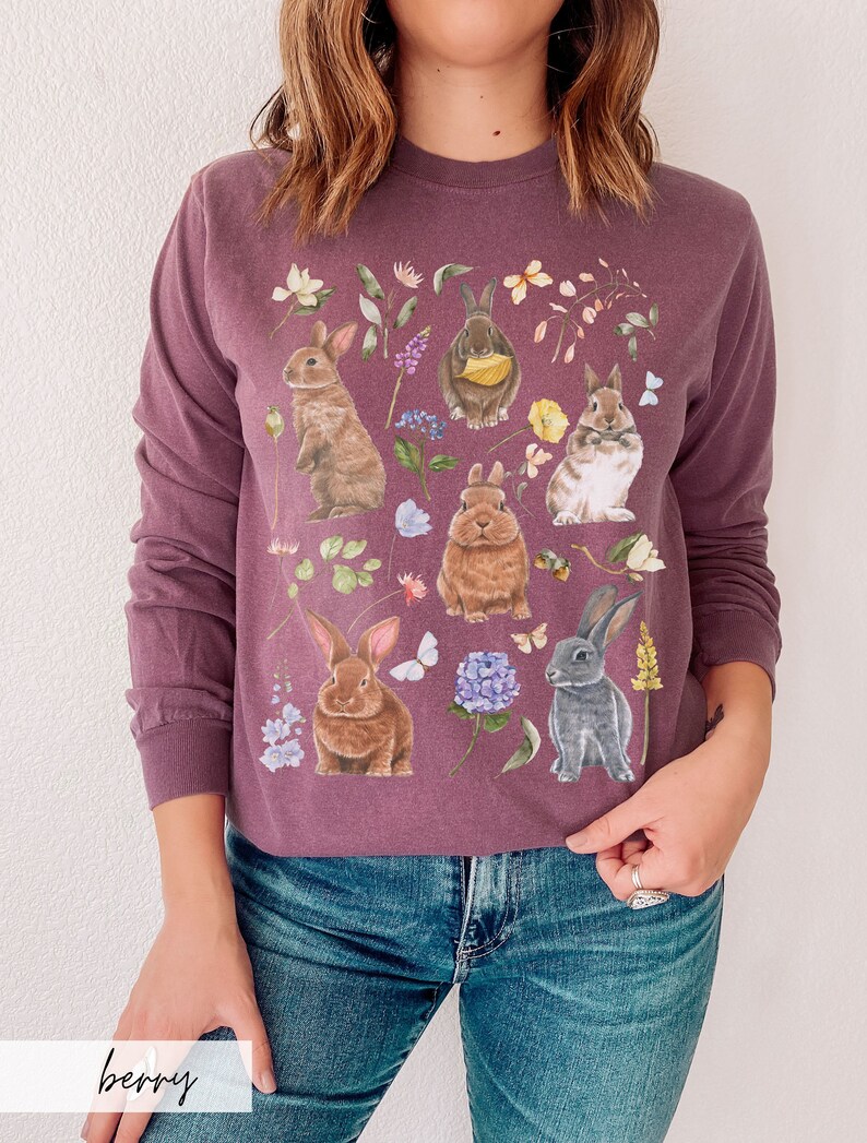 May include: A purple long-sleeved sweatshirt with a floral print featuring multiple bunnies in various poses. The bunnies are brown, white, and gray with pink noses and whiskers. The flowers are in shades of pink, purple, blue, and yellow.