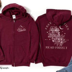 May include: Maroon zip-up hoodie with a white graphic of an open book, moon, and stars on the front. The back features a stack of books, flowers, a crescent moon, and the words "FREE" and "READ FREELY".