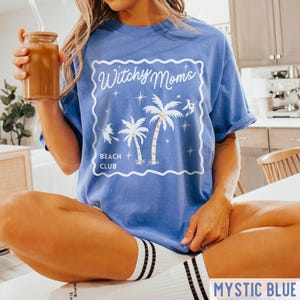 K&ouml;nnte beinhalten: Mystic blaues T-Shirt mit wei&szlig;em Aufdruck "Witchy Moms Beach Club" mit Palmen und Hexen-Silhouetten. Das Shirt hat eine lockere Passform und hochgekrempelte &Auml;rmel. Das Model tr&auml;gt wei&szlig;e Socken mit schwarzen Streifen.