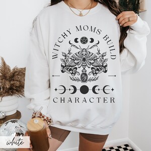 K&ouml;nnte beinhalten: Wei&szlig;es Sweatshirt mit schwarzem Grafikdesign. Das Design beinhaltet eine Motte, Halbmonde und den Text "Witchy Moms Build Character". Das Wort "white" ist unten links aufgedruckt.