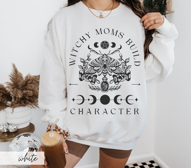 K&ouml;nnte beinhalten: Wei&szlig;es Sweatshirt mit schwarzem Grafikdesign. Das Design beinhaltet eine Motte, Halbmonde und den Text "Witchy Moms Build Character". Das Wort "white" ist unten links aufgedruckt.