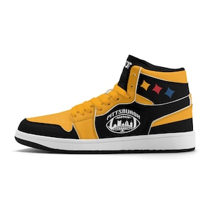 Peut inclure: Basket montante jaune, noire et blanche, avec le mot "PITTSBURGH" et un graphisme de football am&eacute;ricain. La chaussure a un empi&egrave;cement de talon noir avec trois diamants color&eacute;s et une semelle blanche.