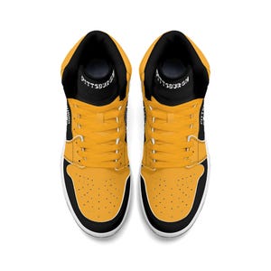 Peut inclure: Paire de baskets montantes, vue de dessus. Les chaussures ont un col et une languette noirs avec le mot "PITTSBURGH" imprim&eacute; sur la languette. Le corps de la chaussure est jaune avec des accents noirs et une semelle blanche.
