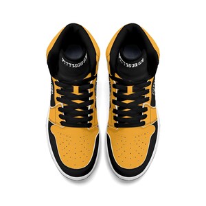 Peut inclure: Paire de baskets montantes, vue de dessus. Les baskets pr&eacute;sentent une palette de couleurs noire et jaune avec des lacets noirs et un col noir. Le mot "PITTSBURGH" est imprim&eacute; sur la languette.