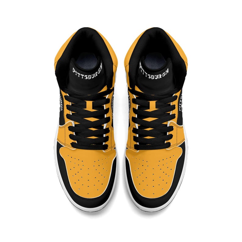 Peut inclure: Paire de baskets montantes, vue de dessus. Les baskets pr&eacute;sentent une palette de couleurs noire et jaune avec des lacets noirs et un col noir. Le mot "PITTSBURGH" est imprim&eacute; sur la languette.