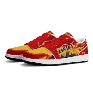 May include: Low-top sneakers featuring a red and yellow color scheme. The side displays the text ESPANA in blue, a dark silhouette of a city skyline, and a small bull icon. The design includes red laces, a white midsole, and a black outsole.