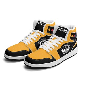 Peut inclure: Paire de baskets montantes jaunes, noires et blanches. Les baskets arborent le mot "Pittsburgh" et un graphisme de football. Semelles et lacets blancs, avec des touches de rouge et d'or. Design sportif.