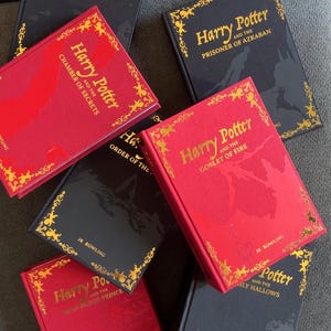 May include: A collection of Harry Potter book editions. The books have red and black covers with gold lettering and decorative borders. Titles include "Harry Potter and the Sorcerer's Stone," "Chamber of Secrets," and "Goblet of Fire."