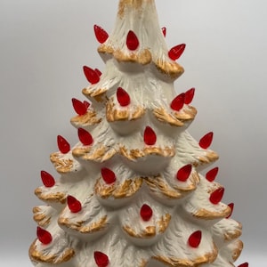 Puede incluir: Un árbol de Navidad de cerámica blanca con bordes pintados en dorado en las ramas. El árbol está decorado con numerosas bombillas pequeñas de plástico rojo translúcido y rematado con una gran estrella roja translúcida.