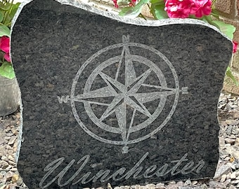 May include: A dark gray granite stone with a compass rose design and the name "Winchester" engraved in a script font. The stone has a natural, irregular shape and is set on a bed of small rocks.