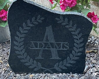 May include: A black granite stone with a carved wreath and the letter A, with the name "ADAMS" written below the letter. The stone is sitting on a bed of small gray rocks.