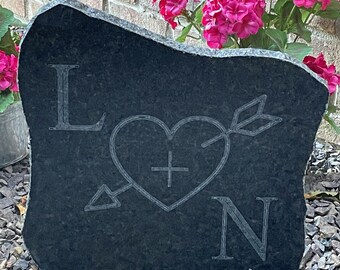 May include: A black heart with an arrow through it and a cross inside, with the letters L and N on either side, carved into a dark gray stone.