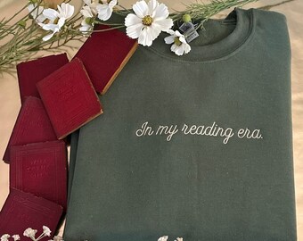 May include: A dark green sweatshirt with the text "In my reading era." embroidered in white on the front. The sweatshirt is surrounded by small red books and white flowers.