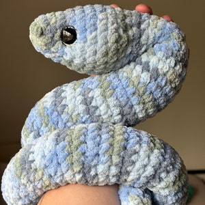 May include: A crocheted blue and green snake plush toy with a single black eye. The snake is curled up in a coil.