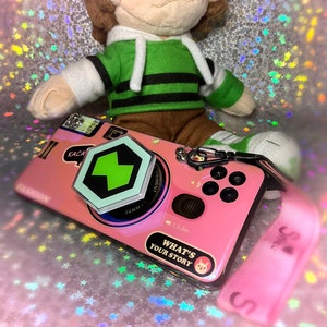 Può includere: Una custodia per telefono rosa con un design a forma di fotocamera e un'impugnatura per telefono esagonale verde. La custodia ha il testo "What's Your Story" e un piccolo adesivo di un gatto. La custodia per telefono è attaccata a un cinturino per telefono rosa con la parola "Smile" su di esso.