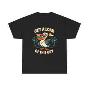 Pode incluir: Camiseta preta com um desenho animado de um pelicano segurando um hambúrguer. O design inclui o texto "GET A LOAD OF THIS GUY" em branco e amarelo, com palmeiras e um sol ao fundo.