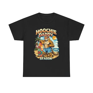 May include: Black t-shirt featuring a cartoon leopard with sunglasses, holding a surfboard on a beach. The shirt has the text "HOOCHIE DADDY SEASON" in a retro style, with a palm tree and sun graphic.