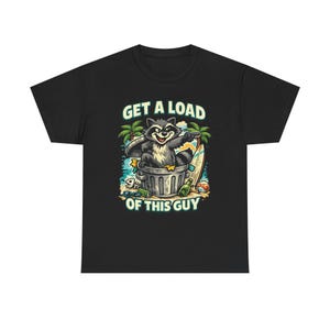 Pode incluir: Camiseta preta com um guaxinim de desenho animado em uma lata de lixo, uma prancha de surf e uma cena de praia. O texto "GET A LOAD OF THIS GUY" é impresso acima e abaixo da ilustração do guaxinim.