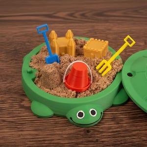 May include: A green turtle-shaped sandbox filled with sand and miniature toys. The set includes a blue shovel, yellow rake, red bucket, and two yellow sandcastles. The lid is also green and shaped like a turtle shell.