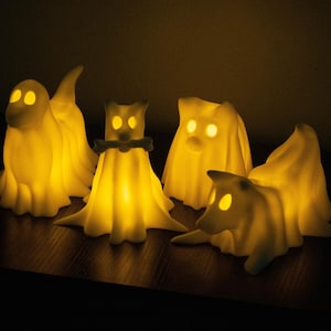 May include: Five glowing white ghost figures with different shapes and sizes. The ghosts are illuminated from within, creating a warm, inviting glow. The ghosts are arranged on a dark wooden surface.