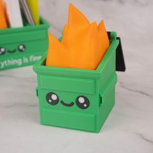 Puede incluir: Organizador de escritorio verde y naranja con forma de contenedor de basura de dibujos animados con fuego en la parte superior. El contenedor de basura tiene una cara sonriente y el texto "Todo est&aacute; bien" en la parte delantera.