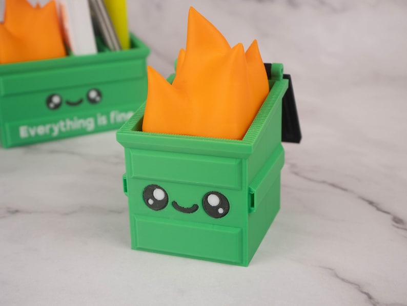 Puede incluir: Organizador de escritorio verde y naranja con forma de contenedor de basura de dibujos animados con fuego en la parte superior. El contenedor de basura tiene una cara sonriente y el texto "Todo est&aacute; bien" en la parte delantera.