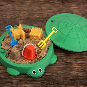 May include: A green turtle-shaped sandbox toy filled with sand and miniature toys. Includes a yellow rake, blue shovel, orange bucket, and yellow sandcastle and block. The lid is off to the side.